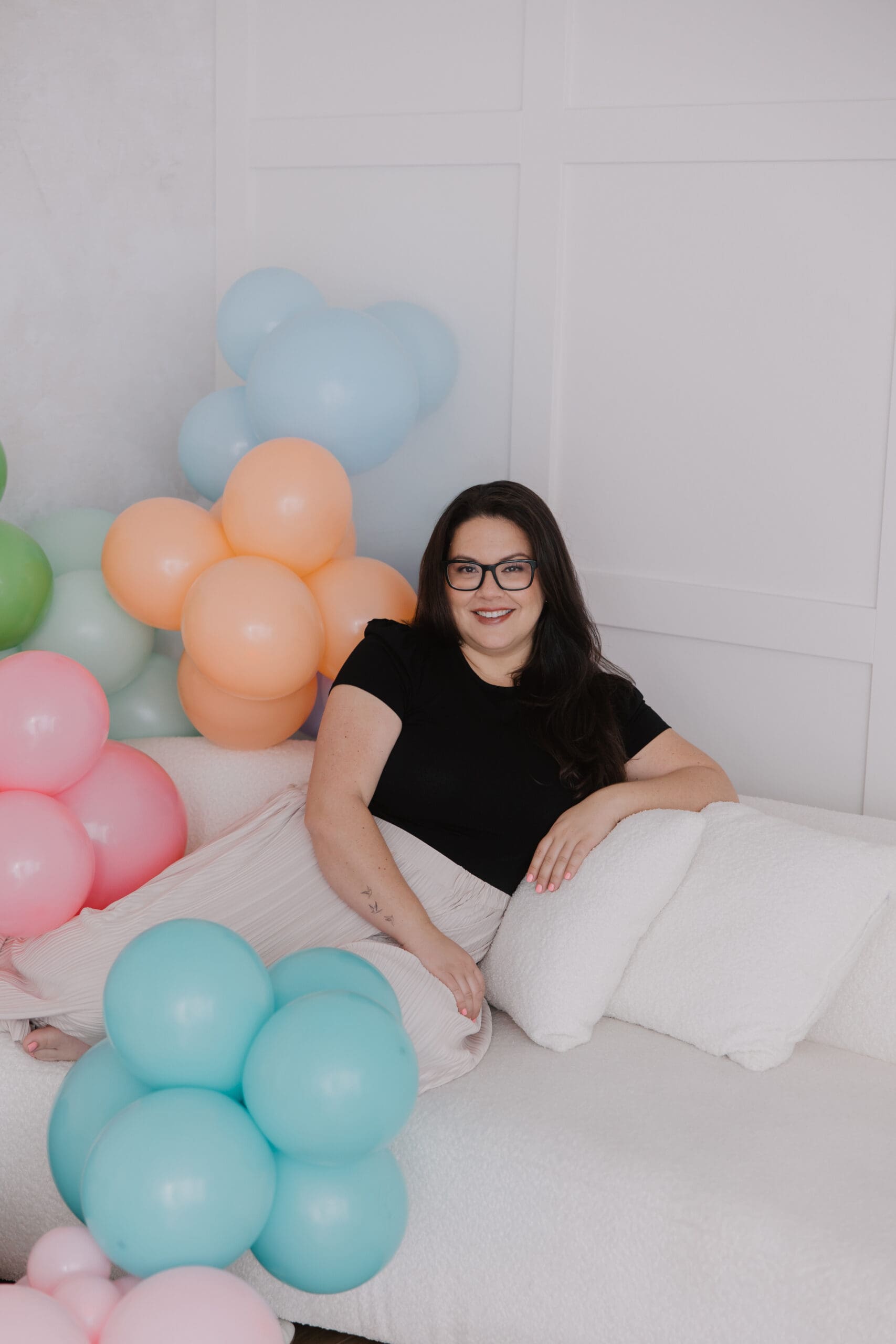 Whitney, founder of The Balloon Bar Co., smiling while seated on a white couch surrounded by pastel balloon clusters in pink, blue, peach, and green during a branding photoshoot.