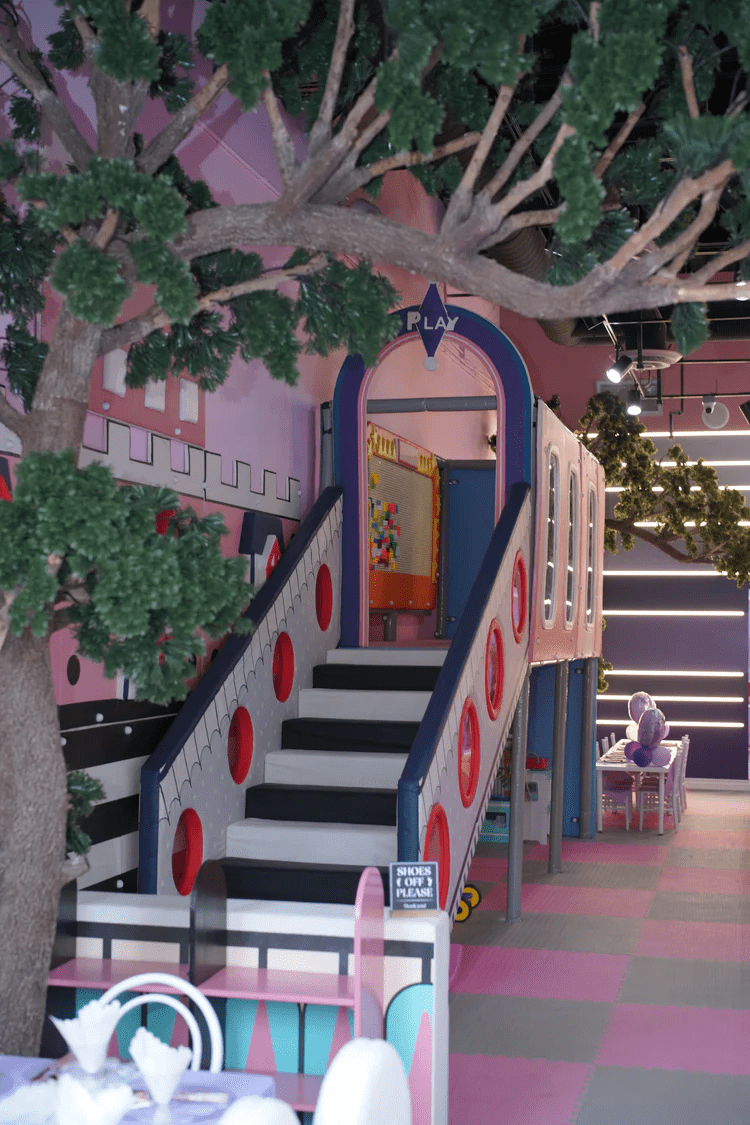 Indoor play space featuring a colorful, castle-themed play structure with stairs leading to an arched entrance labeled “Play,” surrounded by faux trees and soft pastel décor.