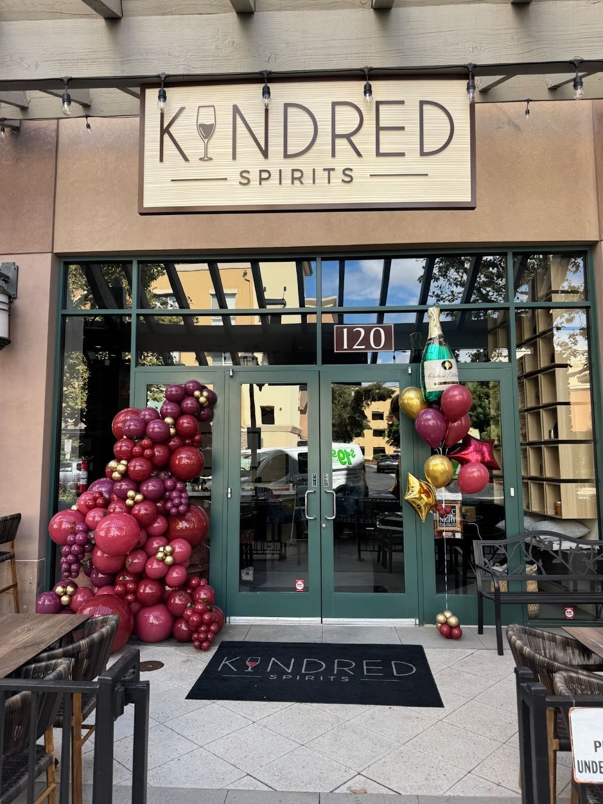 Exterior of Kindred Spirits storefront with green double doors, featuring a burgundy and gold organic balloon installation on the left and a celebratory balloon bouquet with star balloons and a champagne bottle balloon on the right.